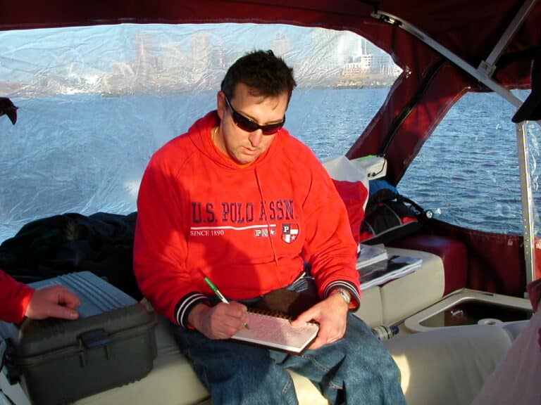 A man wearing sunglasses and a red hoodie with "U.S. Polo Assn." written on it is sitting on a boat. He is writing in a notebook with a green pen. The boat appears to be on a body of water with landscape visible in the background.