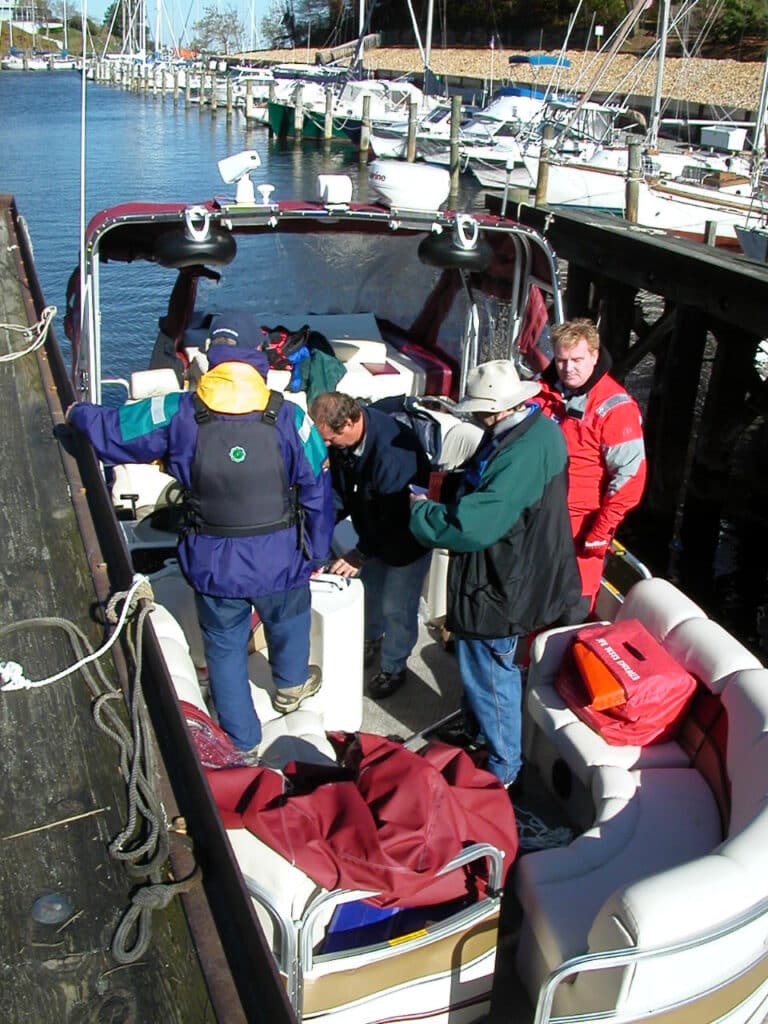 A group of five people on a boat are preparing to set off from a dock. They are dressed in various outdoor jackets and life vests, and appear to be discussing or adjusting something on the boat's deck. Other boats are moored at the dock in the background.