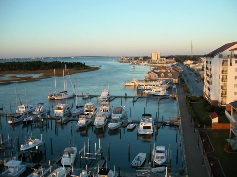 Aerial view of a marina filled with various boats docked at piers. The marina is adjacent to a town with buildings lining the waterway. Beyond the town, the body of water extends towards the horizon with strips of land visible in the distance. The sky is clear.