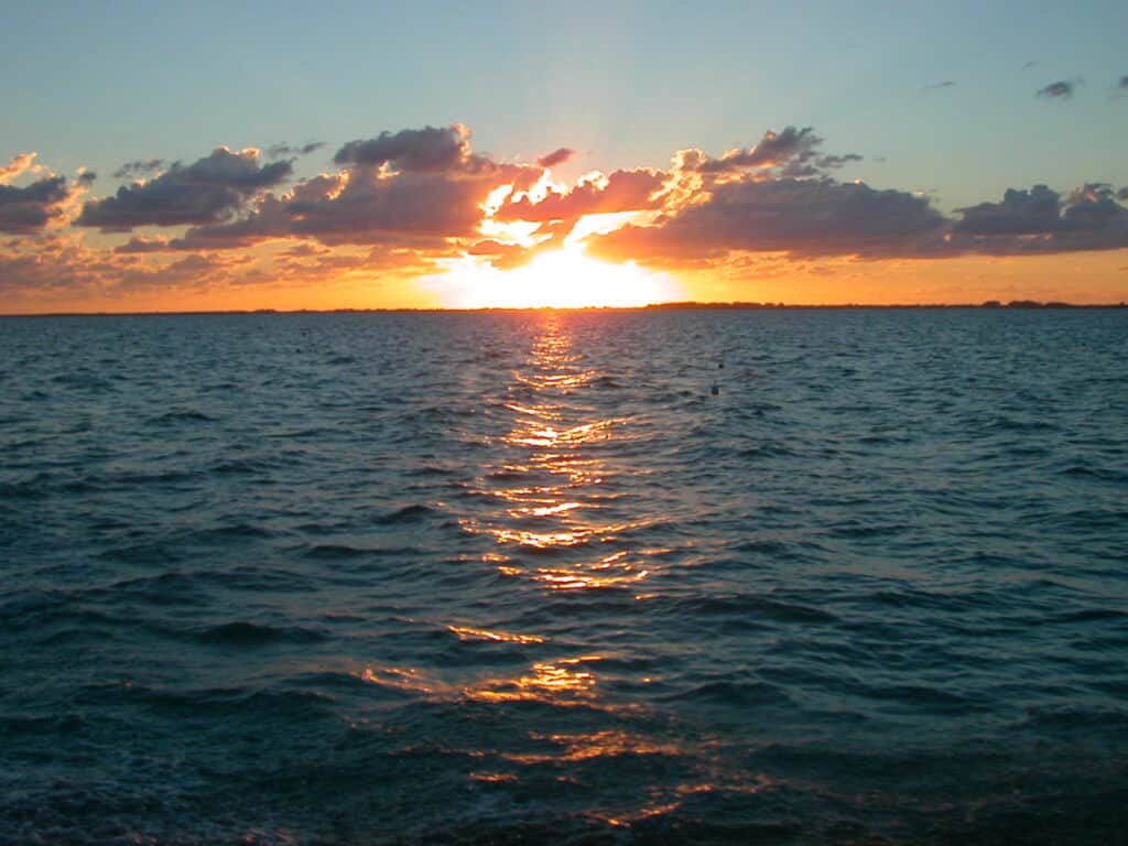 A mesmerizing ocean sunset with the sun setting at the horizon, casting a golden-orange hue across the water. The sky is partly cloudy, with the clouds illuminated by the warm sunset glow, reflecting on the calm sea.