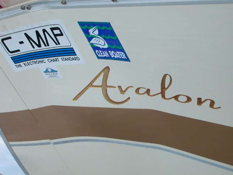 A section of a boat showing three stickers and the name "Avalon." The stickers read "C-MAP, THE ELECTRONIC CHART STANDARD," "CLEAN BOATING," and "BOAT U.S. MEMBER." The boat's exterior features a beige and brown color scheme.