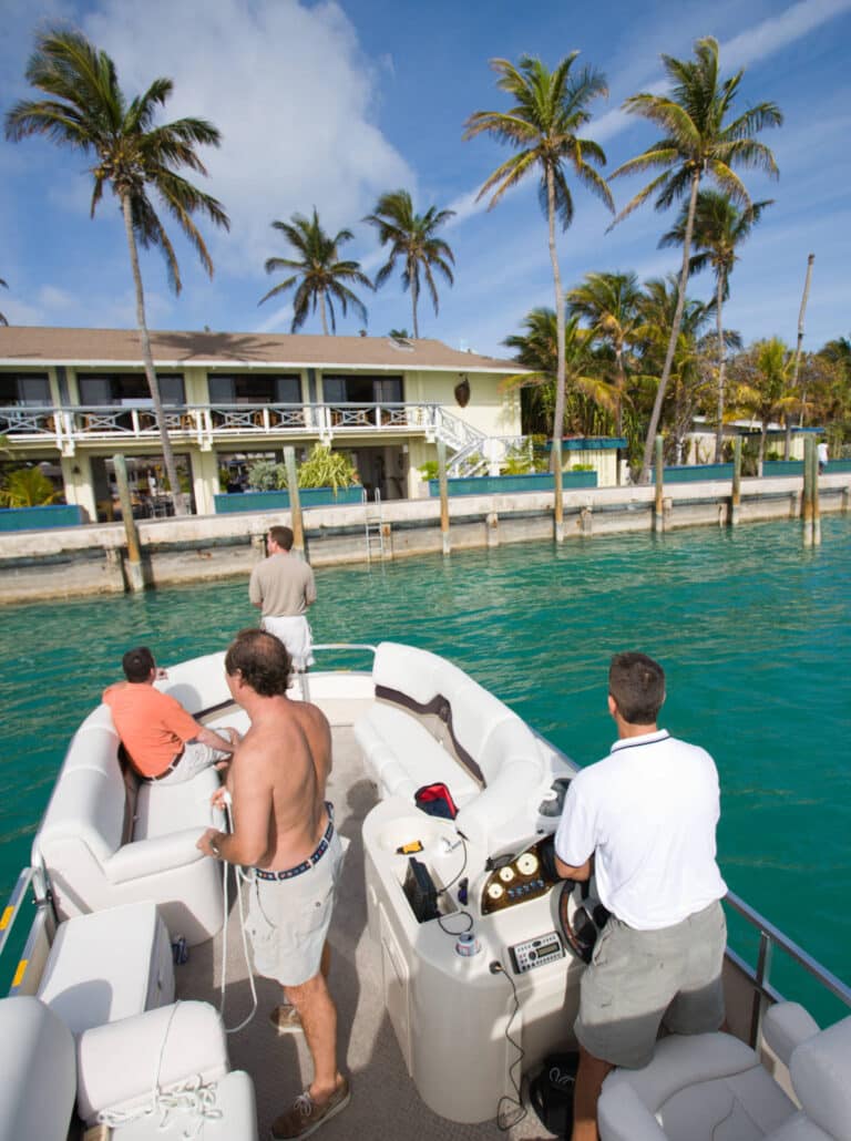 A group of people on a motorboat in turquoise water. The boat is near a dock in front of a tropical house surrounded by palm trees. One person is steering, another is securing a rope, and two others are at the front looking towards the house. The sky is clear.