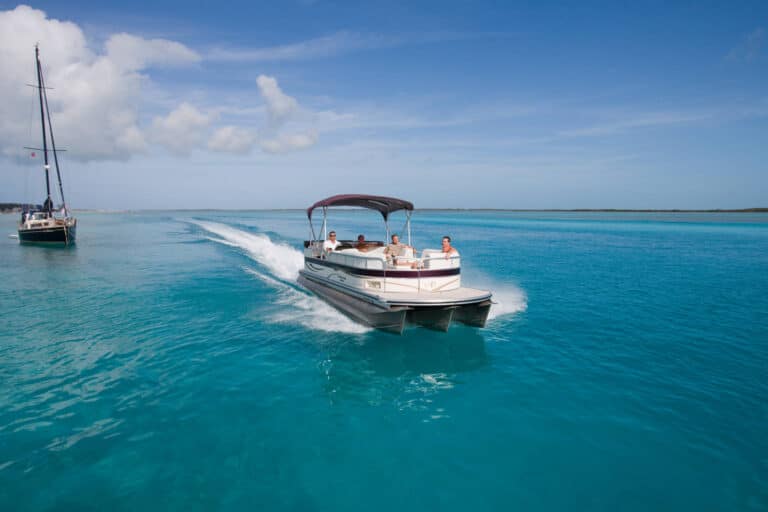 A pontoon boat with a canopy cruises through clear, turquoise water under a blue sky. Several people are on board, enjoying the ride. A distant sailboat is to the left near the horizon. The scene suggests a leisurely day on a calm, tropical sea.