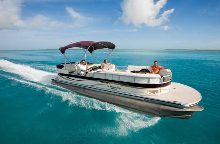 A pontoon boat with a maroon and white canopy cruises on clear blue water. Three people are enjoying the ride, sitting on the boat's white seats, with one person at the helm. The sky is mostly clear with some scattered clouds.