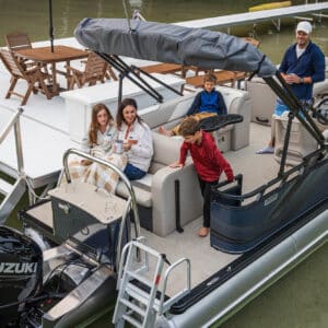 A group of five people, including two adults and three children, are on a Venture Cruise Rear Bench docked next to a pier. The adults are sitting and holding mugs, while the children are positioned around them, one climbing down the back. The boat is equipped with a Suzuki motor.