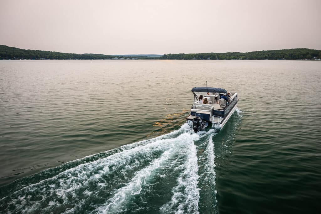 A Venture Cruise Rear Bench with a few passengers is traveling across a calm lake, leaving a white wake behind. The surrounding shoreline is covered with dense green trees under a clear sky, creating a peaceful and serene atmosphere.