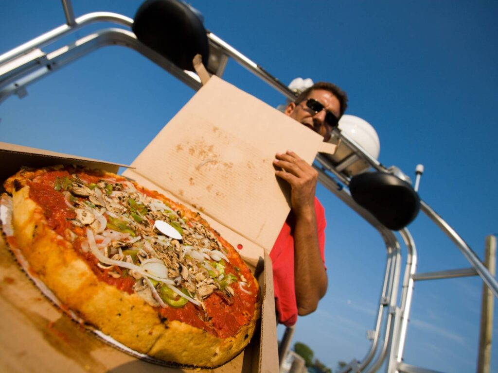 A person wearing sunglasses holds an open cardboard box containing a deep-dish pizza topped with vegetables, including green peppers, onions, and mushrooms. The person is standing outside with a sunny blue sky and metal structures in the background.