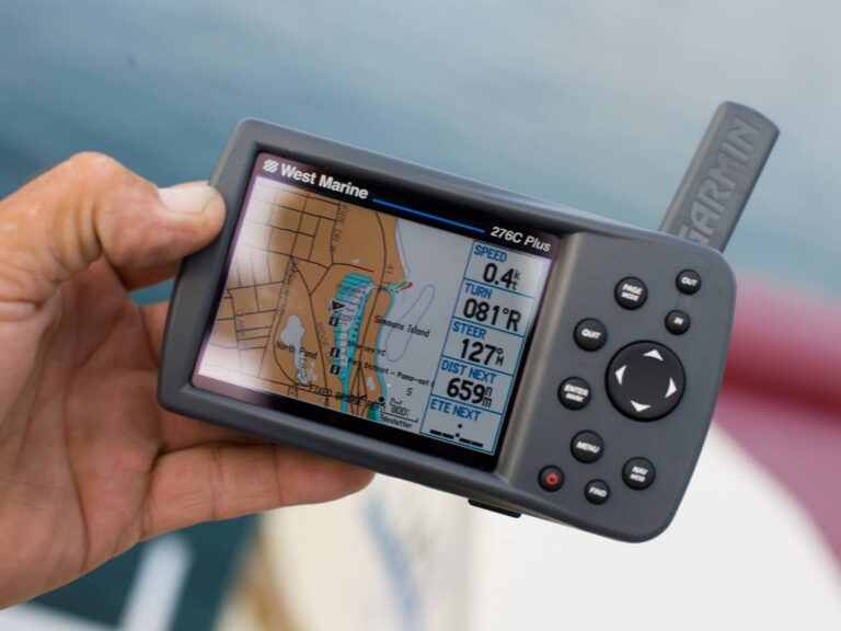 A hand holding a Garmin marine GPS device with a color screen displaying a nautical chart. The screen shows various data including speed, heading, and distance to the next waypoint. The device is black with a keypad on the right side and a visible antenna.