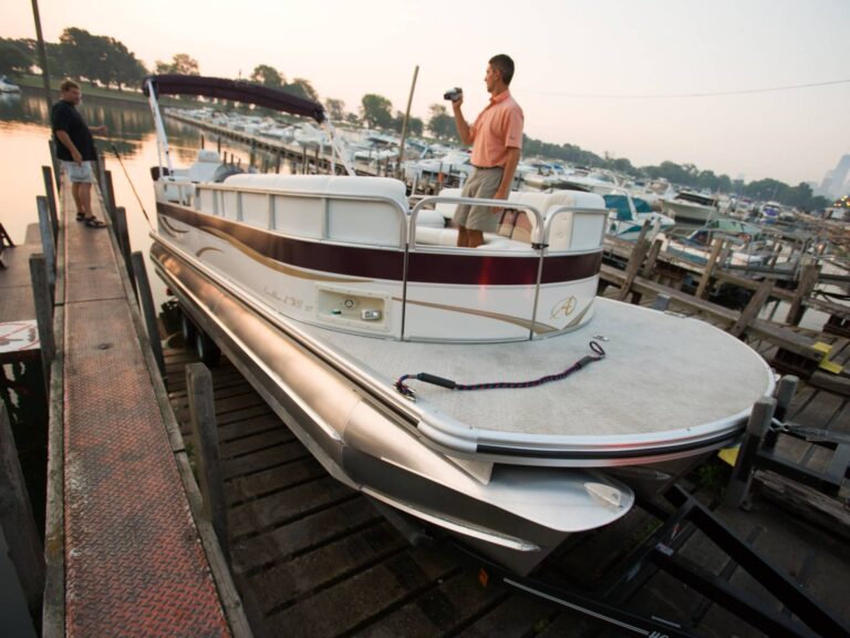 A man stands on a white and maroon pontoon boat docked at a pier, holding a video camera. Another man stands on the dock nearby. The boat is parked at a marina with numerous other boats in the background. The setting appears to be in the early evening.