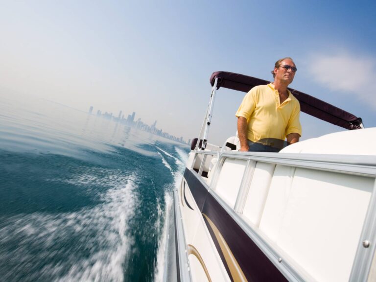 A man wearing sunglasses and a yellow shirt steers a motorboat on calm water. The city skyline is visible in the background under a clear, blue sky. The boat leaves a trail of white wake behind it.