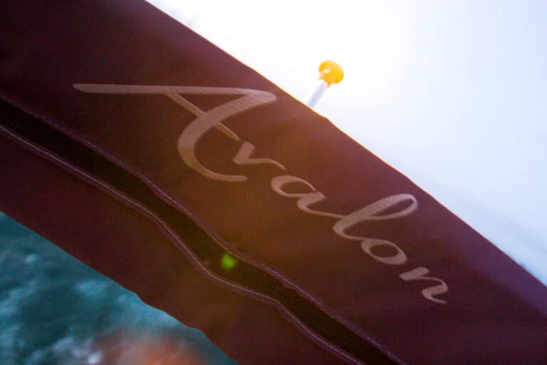 Close-up of a maroon canvas canopy or cover on a boat with the word "Avalon" written on it in cursive. A small part of a yellow light and a blurry background of water and sky are visible.