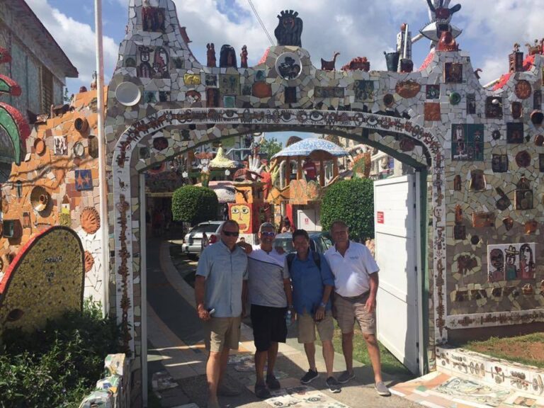 Four men stand together in front of a colorful, mosaic-decorated archway. The intricately designed arch is adorned with various tiles, artwork, and patterns. A street and other artistic structures are visible in the background. The sky is partly cloudy.
