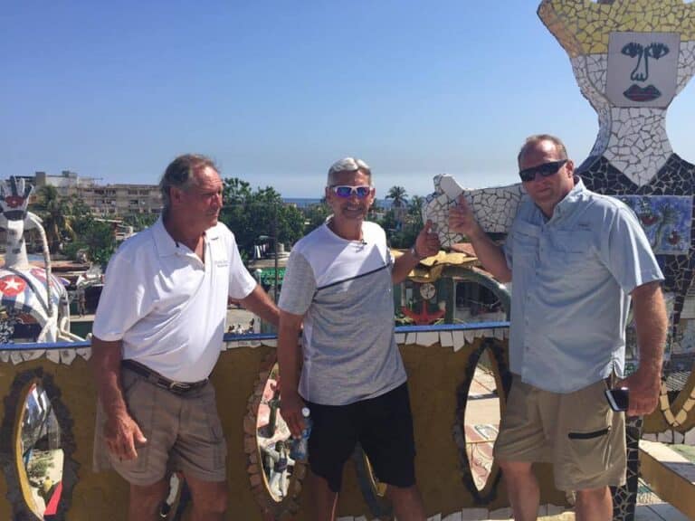 Three men stand together outdoors in front of a colorful mosaic sculpture. The man on the left wears a white polo shirt, the man in the middle wears a light grey shirt, and the man on the right wears a light blue shirt. They are smiling, and the sky is clear.