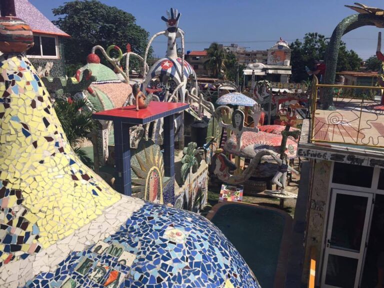A vibrant, mosaic-covered outdoor space features colorful tiles and sculptures, including animals and abstract shapes. The setting has a whimsical, artistic feel with various objects and structures adorned in intricate patterns. Trees and buildings are visible in the background.