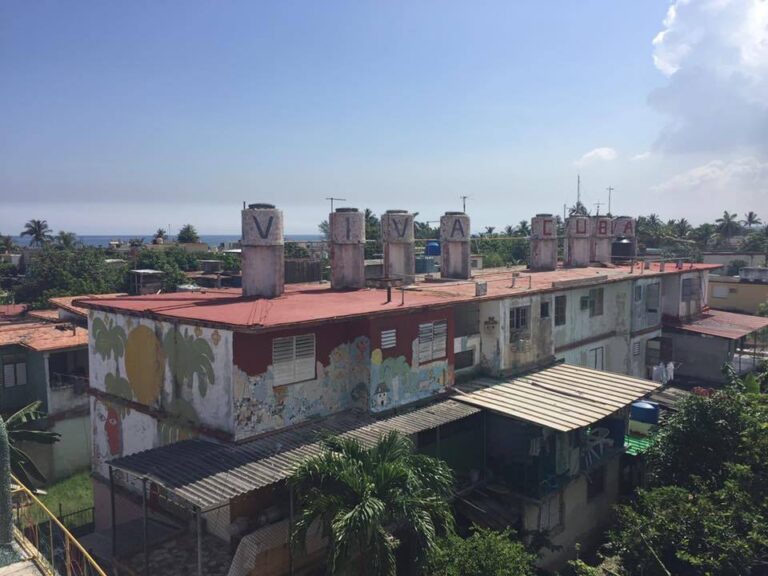 A two-story building with a red roof and weathered exterior in a tropical area, adorned with faded murals. Large letters on the rooftop spell "VIVA CUBA." Palm trees and lush greenery surround the building, and the ocean is visible in the background.