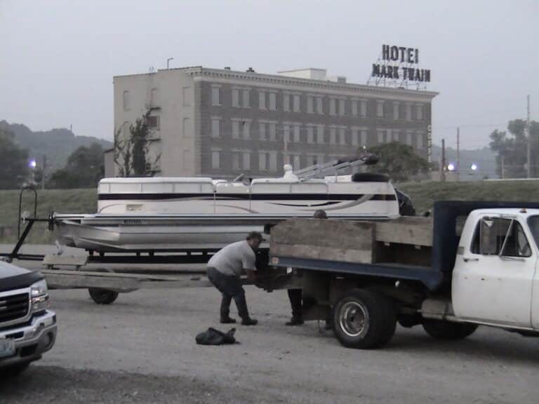 Two men are working on a car with the hood open in a gravel lot. A boat on a trailer is parked behind them. In the background, there is a multi-story brick building with a sign reading "Hotel Mudelein." The sky is overcast, lending the scene a grayish tint.