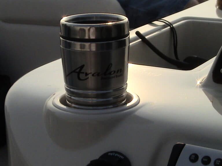 A stainless steel travel mug with the word "Avalon" and "Pontoon Boats" written on it is placed in a cup holder on a boat. The boat's console with control knobs and wires is partially visible in the background.