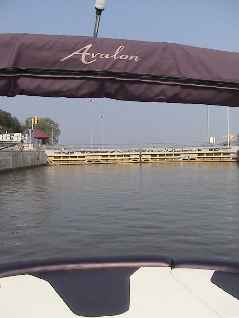A view from a boat named Avalon approaching a canal lock. The boat's canopy is visible at the top. The lock gate is partially open, with calm water leading inside. A small building and trees are seen on the left side, with clear blue skies overhead.