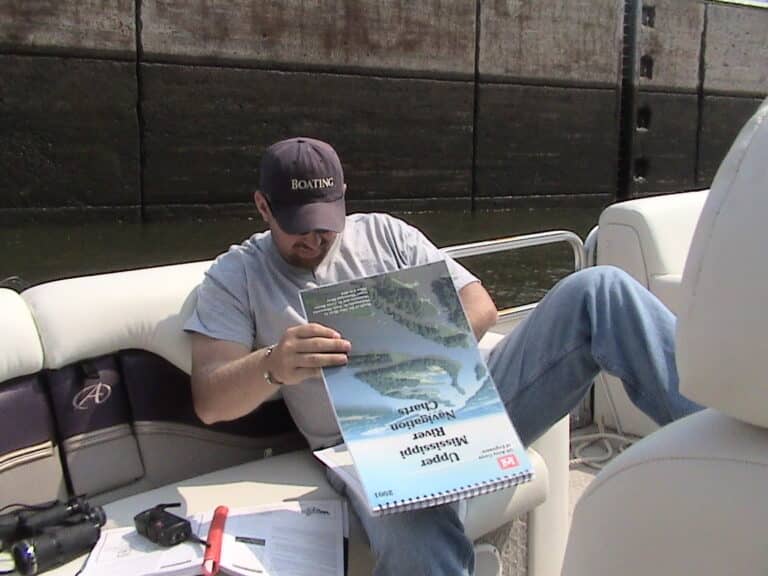 A person wearing a cap labeled "BOATING" and a gray shirt is sitting on a boat. They are reading a large river navigation chart titled "Upper Mississippi River Navigation Charts" with some papers, a pen, and binoculars lying nearby.