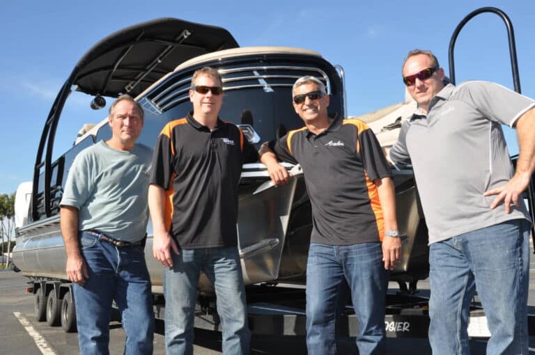 Four men are standing in front of a large boat on a trailer. Three of them are wearing sunglasses, and they are dressed casually in different shades of blue and gray with some in collared shirts. They appear to be smiling and posing for the photo. The sky is clear and blue.