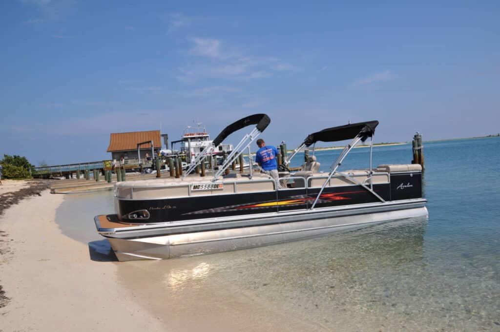 A person stands on a pontoon boat that is beached on a sandy shore. There is a pier with a small building and another boat in the background. The sky is clear and the water is calm.