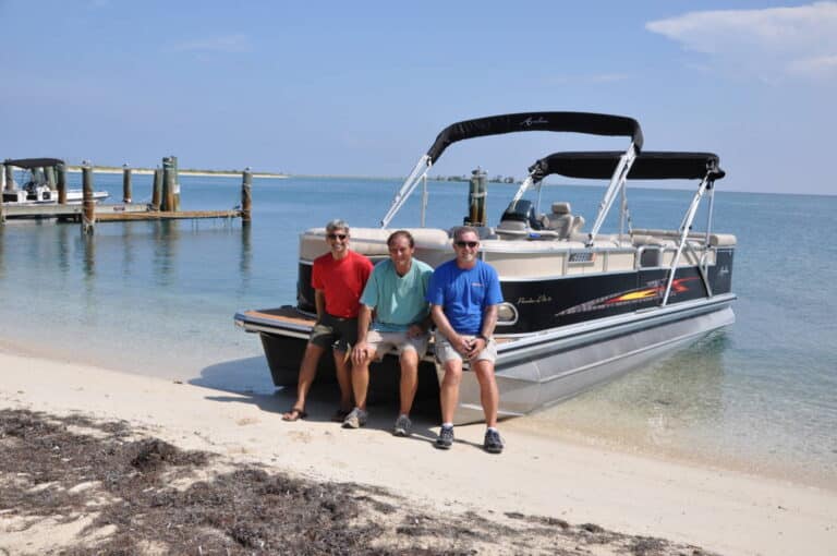 Three men sit on the edge of a pontoon boat pulled up on a sandy beach. The boat has a black canopy and is partially in the water. A dock and another boat are visible in the background under a clear blue sky.