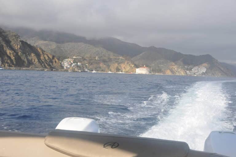 View from a boat in the ocean, showing a wake trailing behind it. The coastline features rocky cliffs with buildings nestled along the base, some structures visible in the distance. Cloudy sky and calm waters set a tranquil scene.