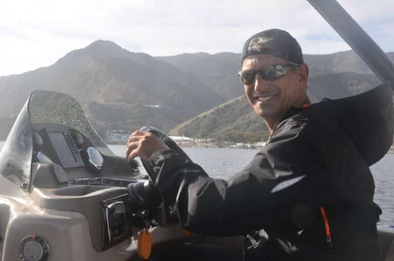 A person wearing a black jacket, sunglasses, and a backward cap is smiling while steering a boat. There are mountains in the background under a slightly overcast sky.