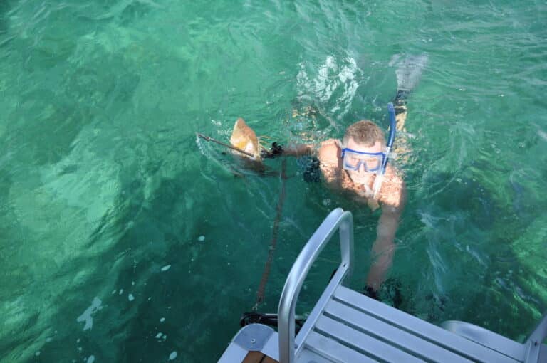 A person wearing snorkeling gear and a blue diving mask is floating in clear, green water next to a boat. They are holding onto a metal pipe connected to the boat's ladder, appearing to climb or hang onto it while looking up at the surface.