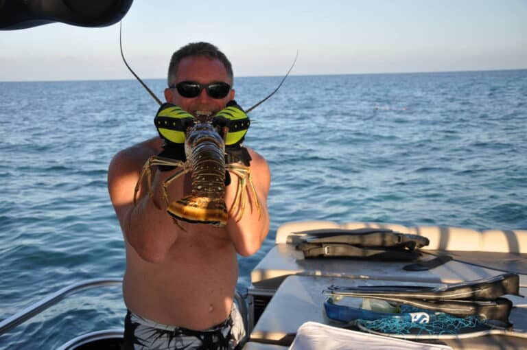 A man wearing sunglasses holds a large lobster up to his face while standing on a boat. The ocean is visible in the background, and the boat's deck has various items, including snorkeling gear and a tackle box.