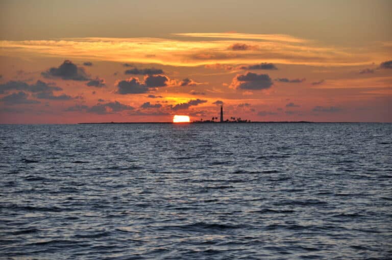 A lighthouse stands tall on a distant island as the sun sets behind it, casting an orange glow across the sky and casting reflections on the calm ocean waters. Scattered clouds add to the picturesque and serene ambiance.