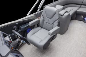 A luxury boat interior featuring a plush, grey captain's chair with armrests, positioned behind a steering wheel and control panel. The area is surrounded by cushioned seating on a light-toned, textured floor, indicating a high-end design.