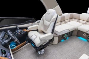 Luxurious boat interior with white and beige leather seating, including a captain's chair with "AVI" on the headrest. The dashboard features a steering wheel and control panel. Ambient blue lighting is visible beneath the seats, and the floor is carpeted.