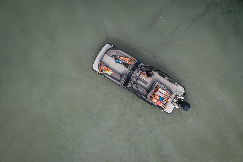 Aerial view of a small boat on water with four people onboard. The individuals are lying down and relaxing, two on each side of the boat. The water is a clear, muted green, and the scene is peaceful with slight ripples around the boat.