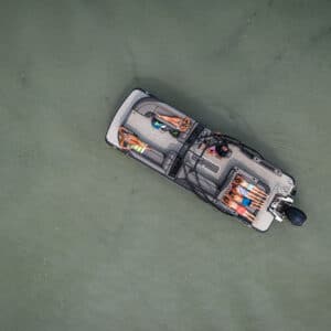 Aerial view of a small boat on water with four people onboard. The individuals are lying down and relaxing, two on each side of the boat. The water is a clear, muted green, and the scene is peaceful with slight ripples around the boat.
