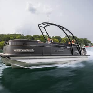 A sleek, black Avalon pontoon boat speeds across a calm lake. Three people are on board, enjoying the ride. The surrounding landscape features lush, green, tree-covered hills under a cloudy sky. The water splashes as the boat moves swiftly.