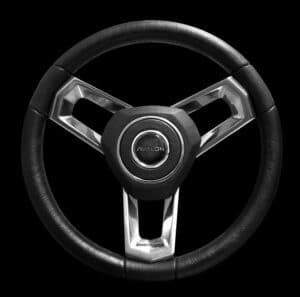A close-up image of a car steering wheel with a sleek, modern design. The three spokes are metallic silver, and the circular center features a logo with "AVALON" on it. The outer rim of the steering wheel is covered in black leather. The background is black.
