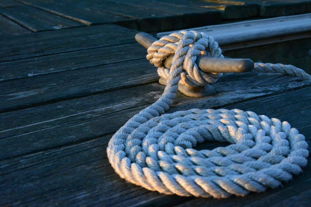 A coiled white rope is tightly secured to a metal cleat on a weathered wooden dock, perfect for docking a pontoon boat. The rope and cleat are illuminated by the warm light of the setting sun, casting soft shadows, while the dock’s wooden planks show signs of age and use.