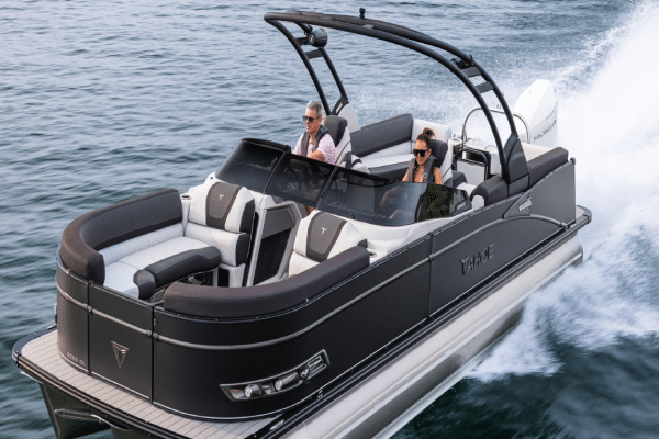 A sleek, modern pontoon boat with two people aboard cruises swiftly over calm waters. The pontoon boat features black and white accents, a spacious seating area, and a dark overhead canopy. Both passengers appear relaxed and are wearing sunglasses.