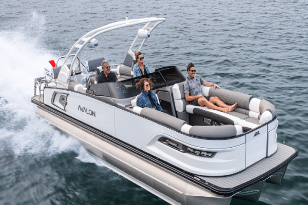 A sleek white and gray Avalon pontoon boat with four passengers cruises swiftly over calm waters. The stylish canopy and cushioned seating add to the charm, with two passengers sitting at the back while the other two are at the front, all enjoying one of the finest pontoon boats around.