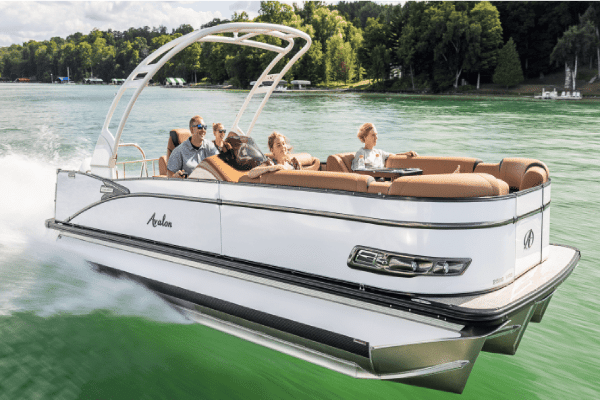 A group of four people enjoys a sunny day on a sleek white pontoon boat for sale with brown seating, cruising swiftly across a calm, green lake surrounded by lush, forested hills. The boat leaves a trail of white water behind.