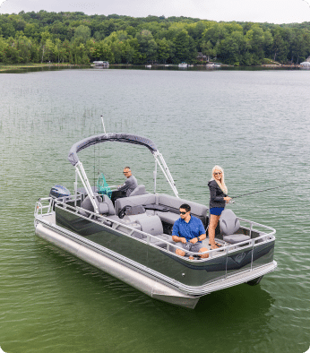 Three people are on a pontoon boat in a calm lake surrounded by forest. One is fishing at the front, while the other two sit and relax under the canopy. The background shows the shoreline with trees and a few houses, making it an idyllic scene for anyone looking at pontoon boats for sale.