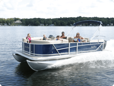 A family enjoys a sunny day on a blue and white luxury pontoon boat in a calm lake. The boat is speeding across the water, creating a wake behind it. Trees and houses are visible on the distant shore under a partly cloudy sky.