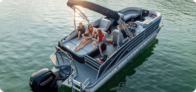 A family of four relaxes on a pontoon boat for sale, floating on a peaceful body of water. Two children sit in the front, while two adults lounge in the back, shaded by a canopy. The boat is anchored by an outboard motor, and the water appears calm and green.