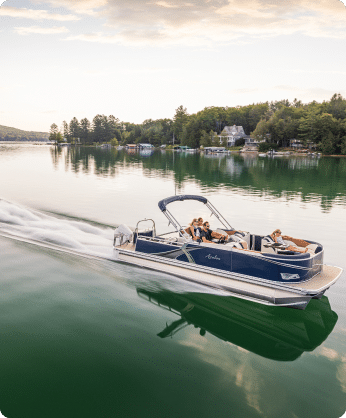 A sleek pontoon boat, one of the many pontoon boats for sale, glides on a reflective lake surrounded by lush greenery and houses in the background. The boat's occupants are seated comfortably, enjoying the serene ride under a cloudy sky. The water's wake trails smoothly behind the boat.