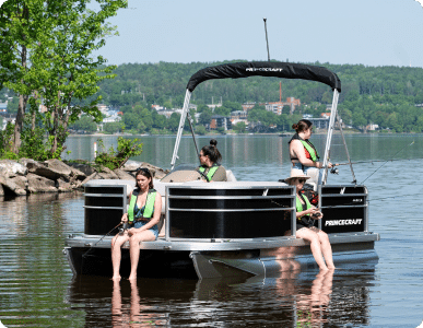 A group of four people fishing on a luxury pontoon boat labeled "Princecraft" in a calm lake. The individuals are wearing life vests, and the boat is near a leafy shoreline with a small town visible in the background across the water.