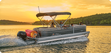 A pontoon boat for sale glides across a calm lake at sunset. The boat has several people on board, some seated and some standing. The sky is ablaze with warm golden hues, and there are lush green trees lining the distant shore.