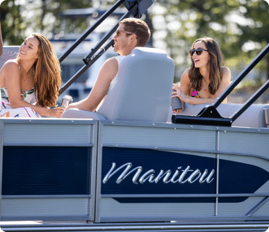 A man and a woman are sitting on a luxury pontoon boat. The woman is wearing sunglasses and holding a drink, smiling while looking at the man who is adjusting the boat's controls. Trees and sunlight are visible in the background.