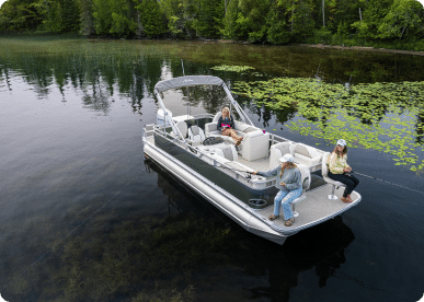 A small pontoon boat for sale floats on a calm lake near a forested shore. Three people, two women and a man, are seated on the boat, with two of them fishing off the sides. Water lilies dot the surface near the shoreline.