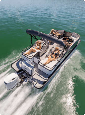 A pontoon boat, one of many pontoon boats for sale, glides quickly across green water. Four people relax onboard, with three lounging on the deck and one person at the controls under a canopy. The boat creates a small white wake as it moves forward.